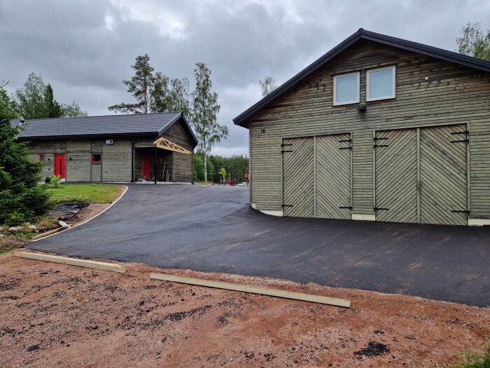 Nyasfalterad garageuppfart med två trähus, den ena med röda dörrar och fönster.orrar i bakgrunden och asfalten omsluter gräsytan.