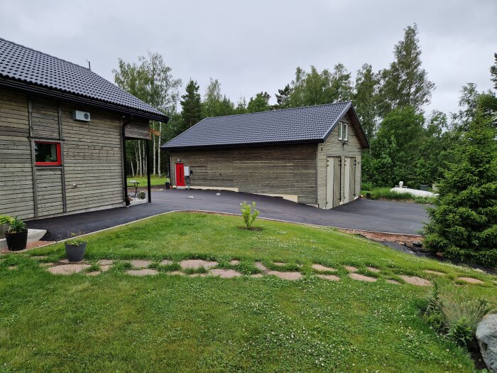 Asfalterad gårdsplan med träbyggnader och grön gräsmatta i förgrunden, omgiven av träd och växtlighet.