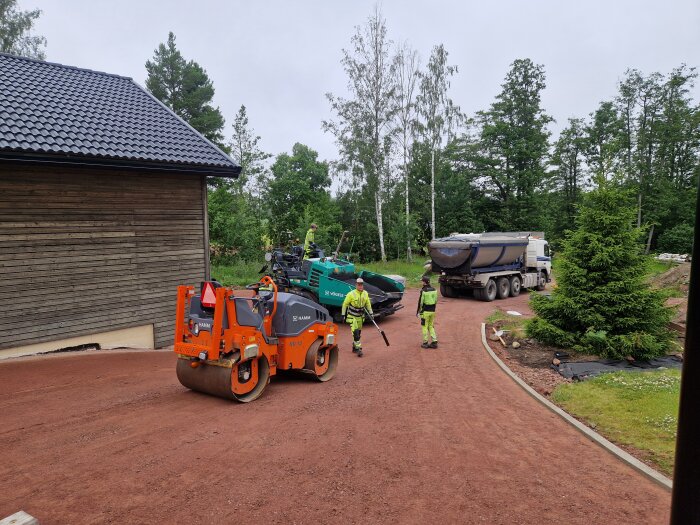 Asfalteringsarbete med en vält, lastbil och två arbetare vid en grusplan nära ett hus och skog.