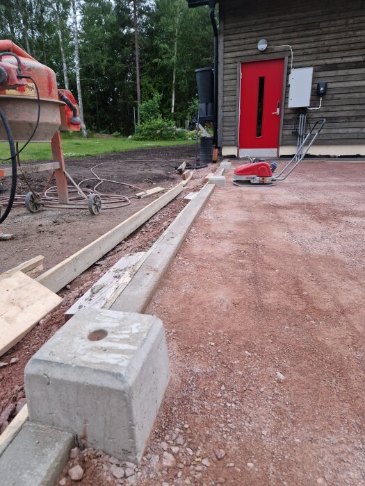 Betongfundament och formarbete nära en träbyggnad med röd dörr. En betongblandare och markberedning ses i bildens framkant.