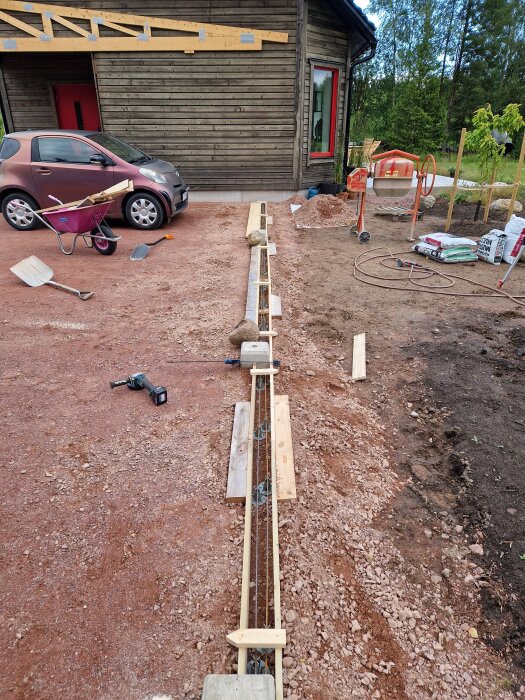 Byggarbetsplats med formar för betonggjutning nära ett hus; verktyg, skottkärra och blandare ses i bakgrunden.