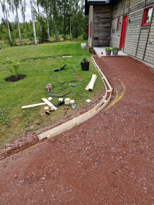 Byggarbetsplats med pågående formarbete för gjutning av stödklack vid en uppfart intill ett hus. Träplankor och verktyg syns på marken.