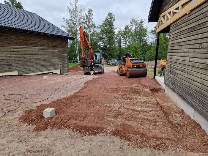 Grävmaskin och vält på grusuppfart mellan två träbyggnader, förbereder underlag för asfaltering. Vitt fundament syns på marken.