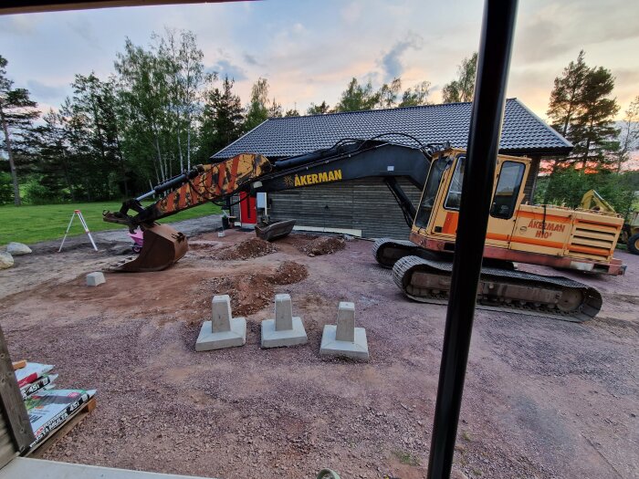 Grävmaskin står intill ett hus på en grusig gårdsplan, omgiven av några betongblock och jordhögar, med träd och gräsmatta i bakgrunden.