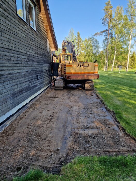 Grävmaskin vid sidan av en träfasad gräver bort ogräs och förbereder området för nya projekt. Gräsyta syns i bakgrunden med träd i fjärran.
