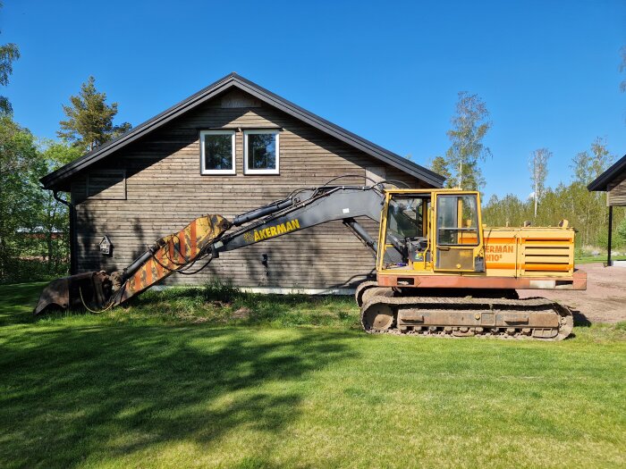 En gul grävmaskin från märket Åkerman står bredvid ett träfärgat hus med grön gräsmatta runt om under en klarblå himmel.