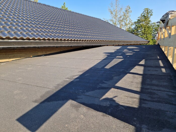 Svart takpapp täcker ett nylagt tak vid en carport, med skuggor från takstolarna och ett traditionellt tak i bakgrunden under en klar himmel.