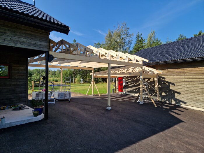 Trästruktur för carport under uppbyggnad, med takstolar och stödjande pelare. Omges av grönska och närliggande byggnader med röda och svarta tak.