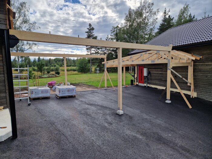 Konstruktion av en carport pågår, stödbalkar och takstolar är monterade. Träbalkar över en asfalterad yta nära ett hus med rött dörr.