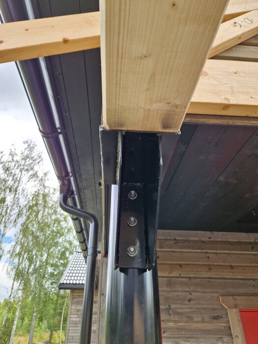 Svart stålstolpe med fastskruvad träbalk och tre bultar vid ett husbygge, synlig ränna och brädvägg i bakgrunden.