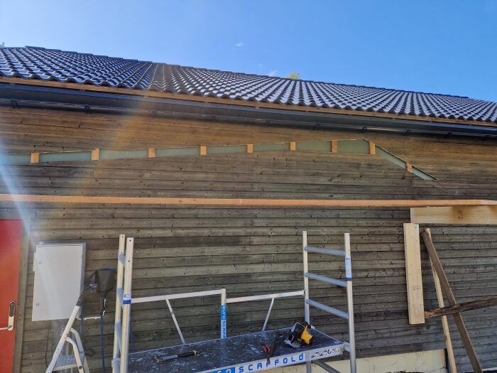 Pågående carportbygge med synliga träreglar och verktyg, en röd dörr, samt en stege framför en trävägg under ett tak.