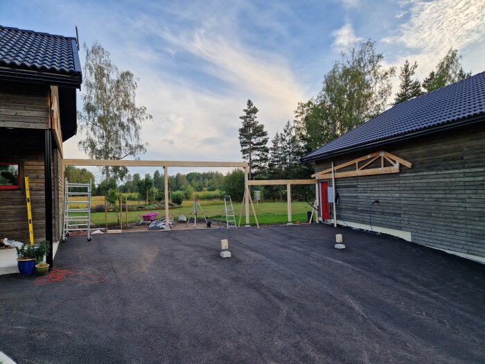 Byggnation av carport: Del av stomme står klar med verktyg och stegar runtomkring. Två hus med tegeltak syns på vardera sida.