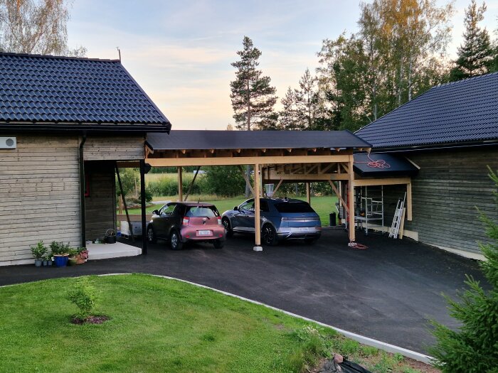 Carport under byggnation med två bilar parkerade, omgiven av trähus och grönska, synligt med råspont och takpapp på taket.