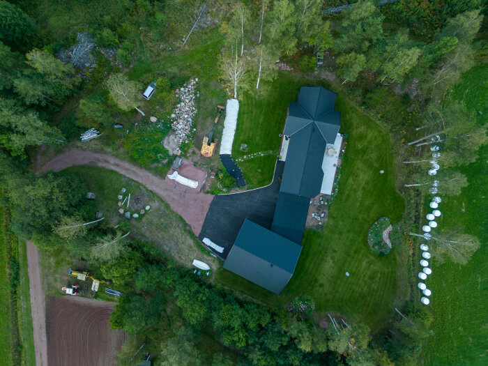 Flygbild av en fastighet med hus, trädgård och skogsomgivning. Gräsmatta omger byggnaderna, och vägar och stigar syns i bakgrunden.
