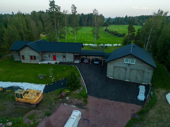 Flygbild av ett hus med carport, stor gårdsplan och trädgård. En grävskopa syns till vänster, omgiven av grönska och skog i bakgrunden.