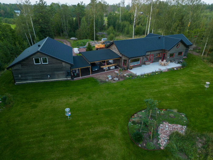 Flygfoto av hus med nybyggd carport och rymlig bakgård med grön gräsmatta och planteringar, omgiven av skog.