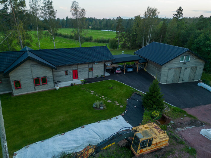 Vy över ett hus med rött dörr- och fönsterram, carport och garage. Framför huset syns gräsmatta och en grävmaskin i förgrunden.