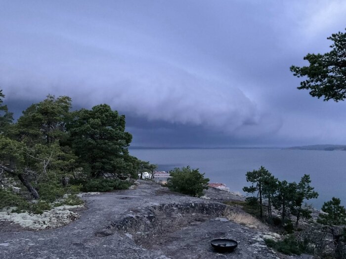Dramatiska mörka moln rullar in över en sjö och landskap medan vinden blåser. Träd syns på klippor i förgrunden.