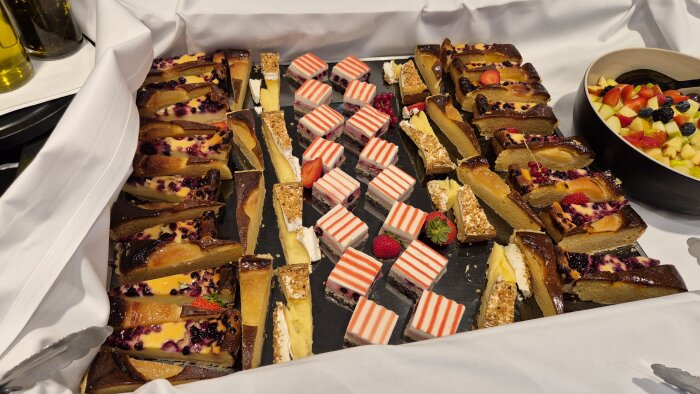 Olika desserter, inklusive bärkakor, randiga tårtbitar och fruktsallad i skål, serverade på en bricka.