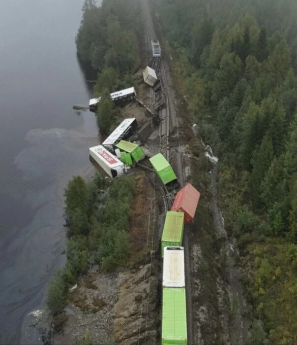 Tågolycka med avspårade godsvagnar i skogsmiljö vid vatten.