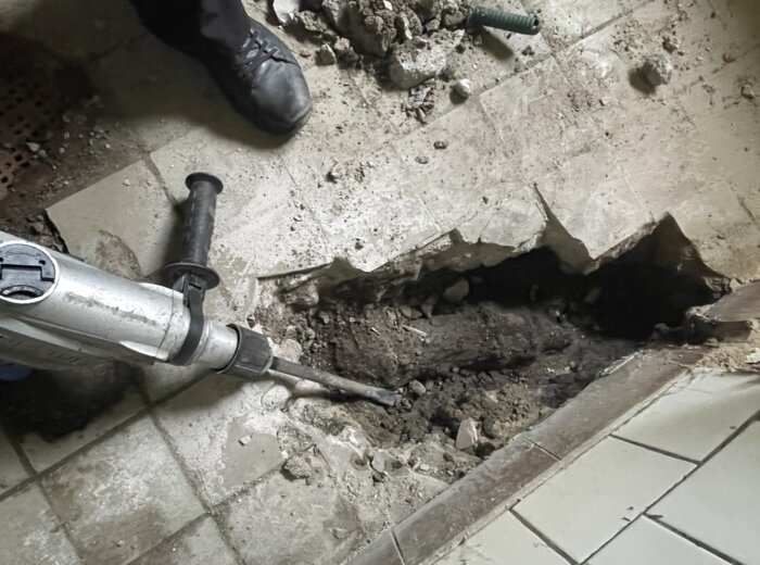 En bilningshammare används för att bryta upp ett hål i ett kaklat golv, med verktygets spets riktad mot ett område med synlig smuts och grus.