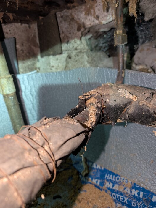 Gamla rör med skadad isolering i en krypgrund, delvis exponerade. Skada troligen orsakad av gnagare. Bakgrund med isoleringsmaterial och stenväggar.