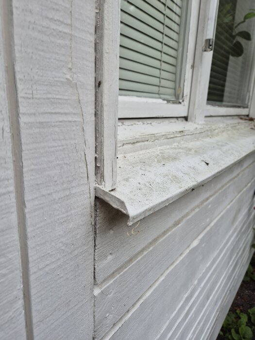 Gammalt vitmålat fönster och fönsterbleck på ett hus, med viss sliten yta och persienner synliga bakom glaset.