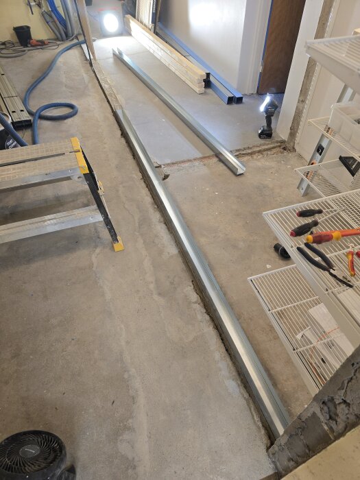 Byggarbetsplats med stålprofiler placerade på ett betonggolv, stege och verktyg synliga. Belysning från arbetslampor.