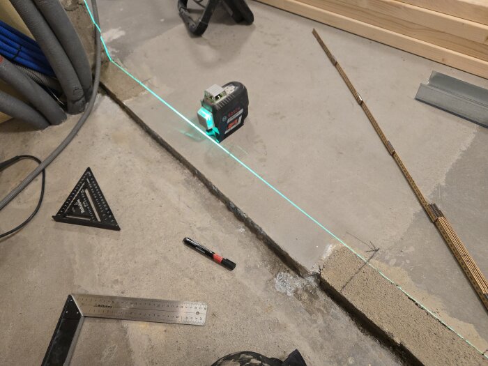 Grön laserlinje på golvet för att mäta och sätta upp stålreglar, omgiven av verktyg som tumstock, vinkelhake och markörpenna.