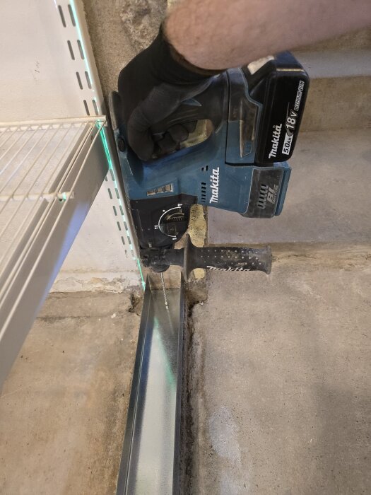 En person använder en elektrisk borrmaskin för att fästa en stålregel mot golvet, med en grön laserlinje som guidning.