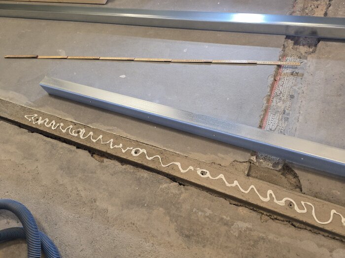 Stålreglar och fogmassa på golvet, en del av byggprocessen för att regla upp väggar på ett betonggolv. Mätsticka och dammsugarslang syns också.