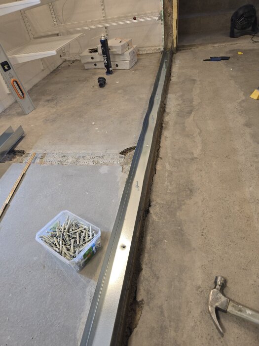 Stålregel monterad på betonggolv med spikplugg. Hammare och låda med pluggar synliga i bild. Hyllplan och tapetrulle i bakgrunden.