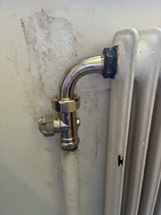 Radiator med nyinstallerad ventil och böjt kopparrör, trångt utrymme mot vitmålad vägg, spår av tidigare reparationer synliga.