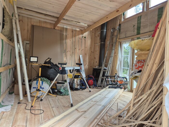 Interiör av ett pågående byggprojekt med verktyg, stege, byggmaterial och en såg på arbetsbockar. Väggen och golvet är av trä.