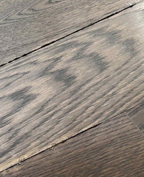 Närbild av ett trägolv med slitna ytor och mellanrum mellan plankorna där något material verkar komma upp. Golvet har ljusa partier och ojämn slipning.