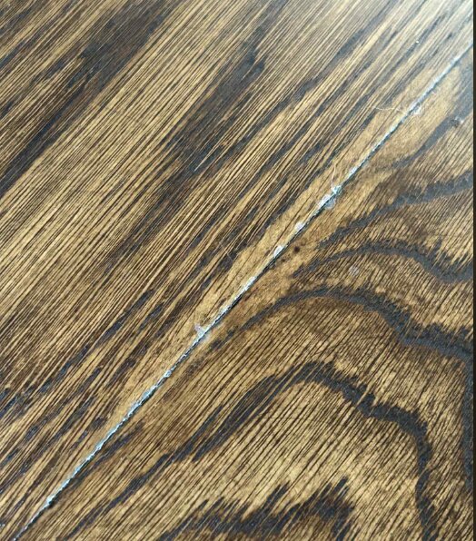 Närbild på ett brunt trägolv med ljusa partier och något material som tränger upp mellan plankorna.