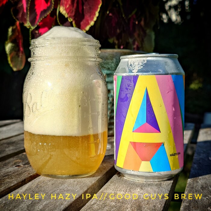 Ölburk och glasburk fylld med Hayley Hazy IPA på ett träbord, omgiven av växtlighet i bakgrunden.