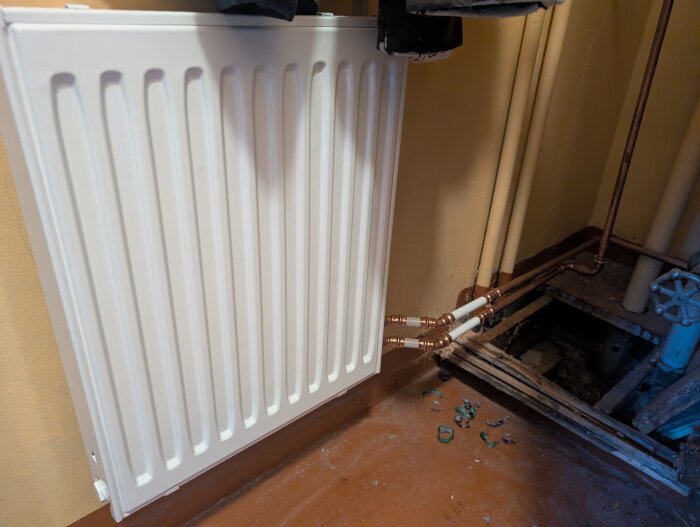 Radiator ansluten med kopparrör, monterad mot vägg med blandarfäste och väggbrickor, golvets öppning synlig.