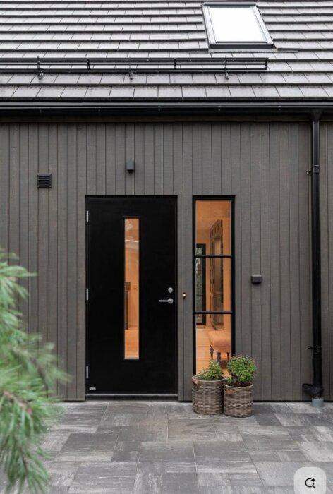 Modern fasad i dämpad svart färg, svart dörr och stora fönster. Två korgar med växter framför entrén. Skiffertak och stenbelagd gång.