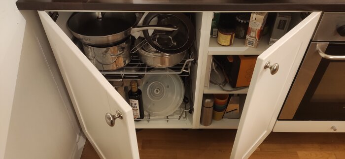 Öppet köksskåp med kastruller och stekpannor i vänstra delen, samt olivolja och köksredskap i högra delen.