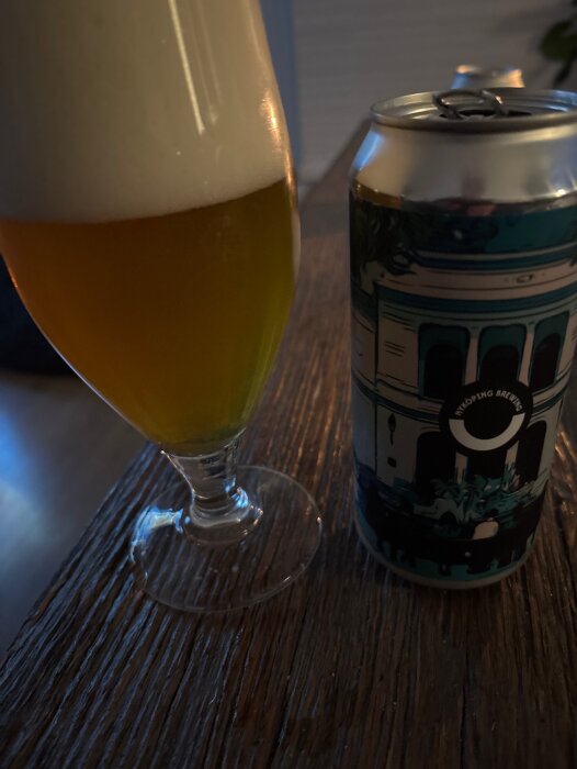 Ett glas med skummande öl bredvid en burk från Nyköping Brewery, fotograferat på ett träbord.
