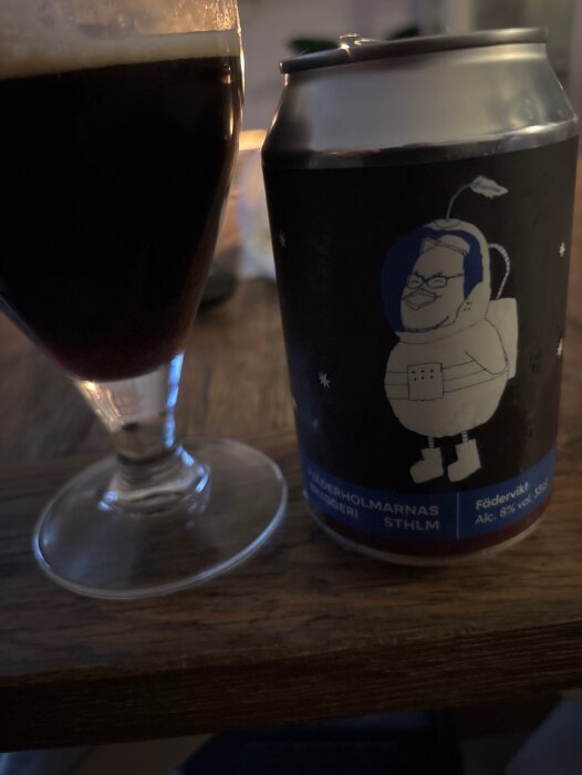 En burk Fjäderholmarnas bryggeri öl med etikettillustration och ett fyllt glas med mörk dryck på ett träbord.