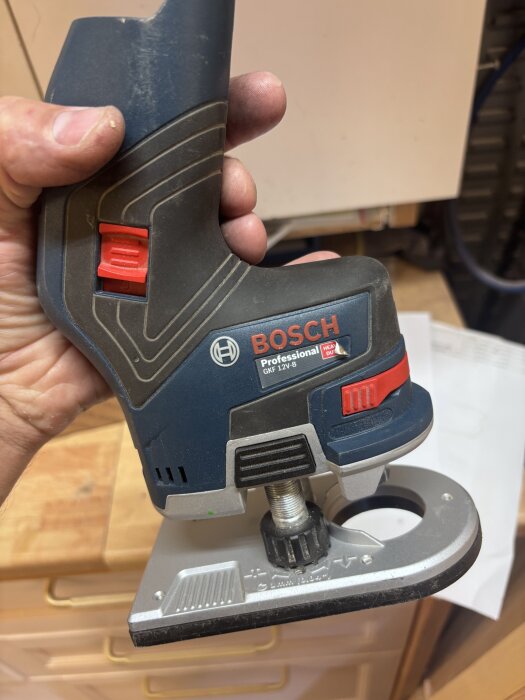 Bosch GKF 12V kantfräs i en hand, svart och röd design, visas på en arbetsbänk i verkstadsmiljö.