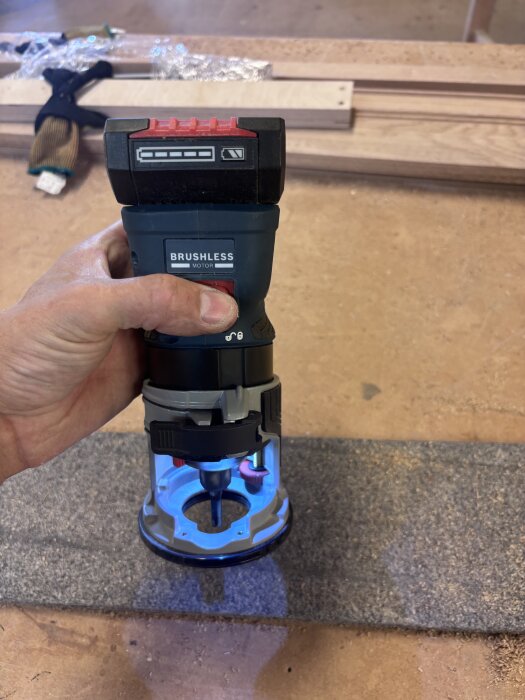 En hand håller en Bosch kantfräs GKF 18 V med en blå lysande lampa, placerad över ett mörkt trästycke på en arbetsbänk.