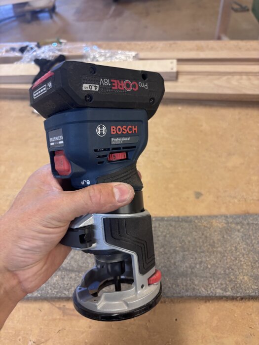Hand holding a Bosch GKF 18V cordless router with a CORE 18V battery, inside a workshop setting.