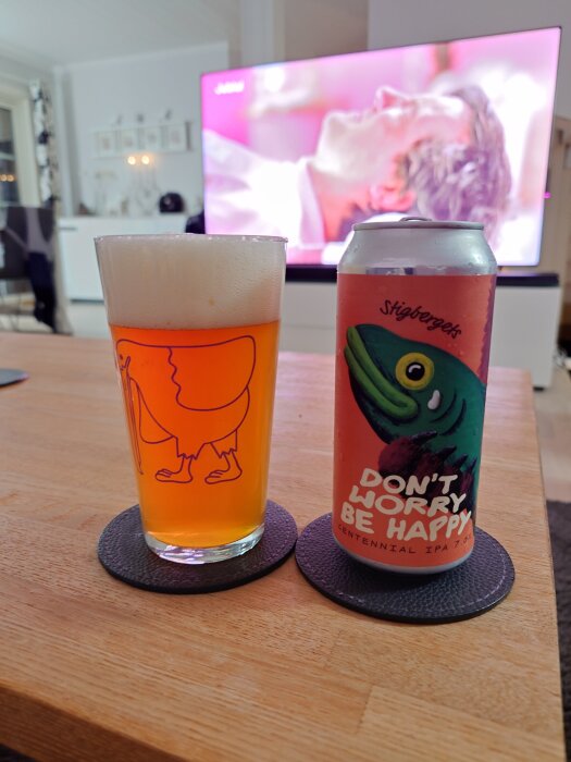 En ölglas och en burk "Don't worry Be happy" IPA på ett bord framför en TV.