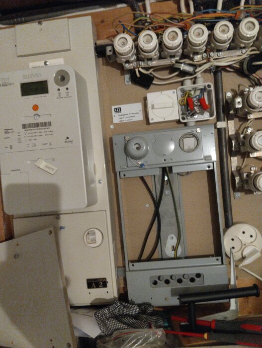 Elcentral med kablar, brytare och äldre mätartavla. En vit vridströmbrytare syns ovanför en gammal mätartavla. Verktyg finns på golvet.