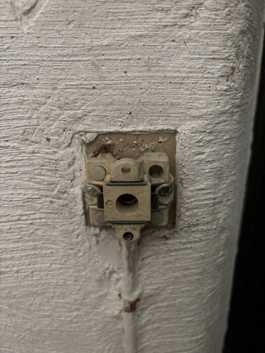 Gammal ljusströmbrytare utan lock monterad på en vitmålad vägg, med synliga skruvar och kablar.