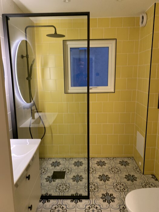 Modernt badrum med gult kakel och svart duschvägg, dekorativa golvplattor i grått och vitt, stor rund spegel med belysning.