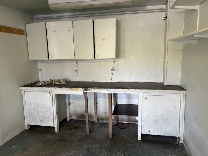 Slitet arbetsbord och fyra vita väggskåp i gammalt garage, med rester av färg och smuts.